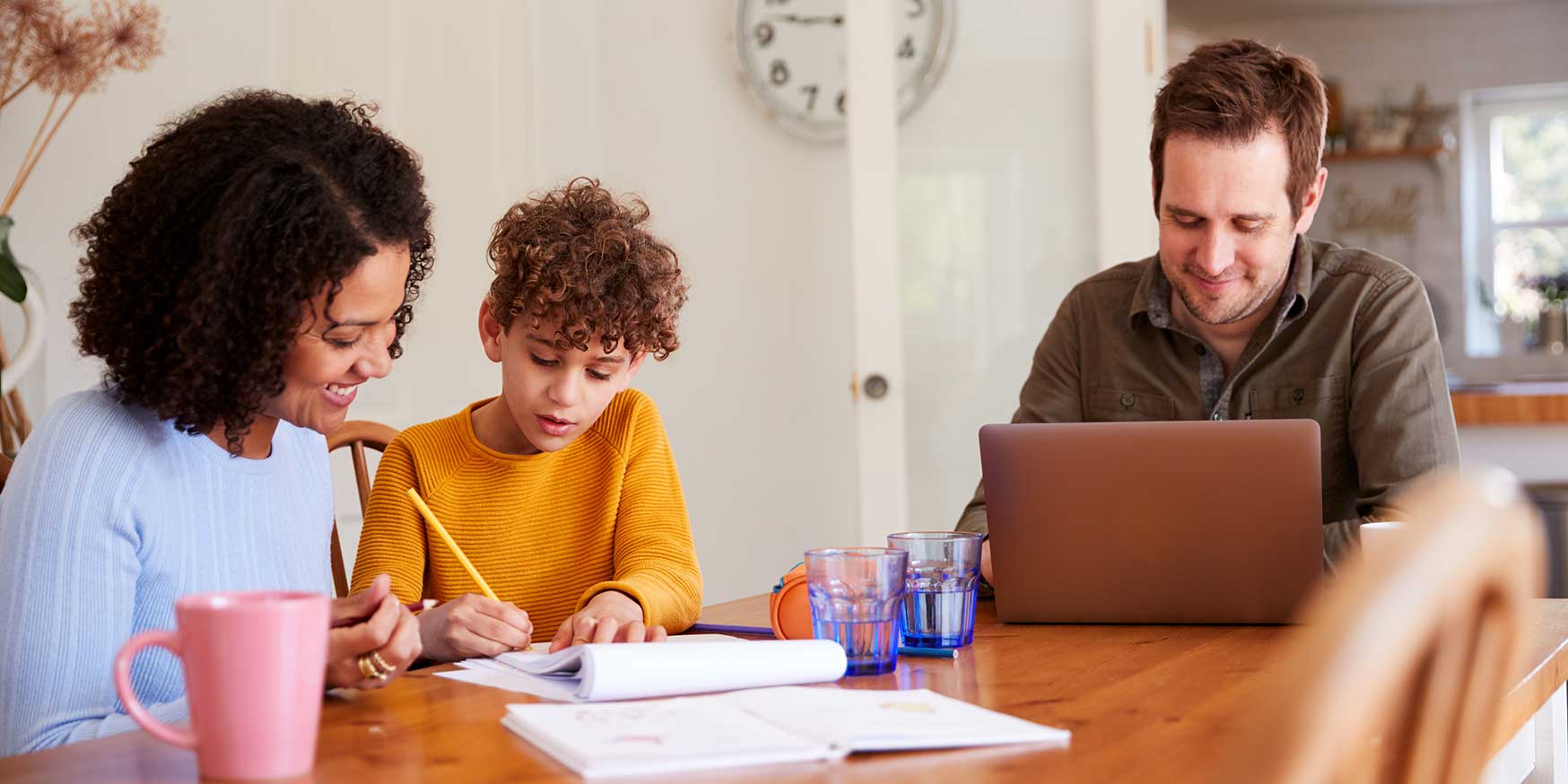 Father Works On Laptop As Mother Helps Son With Homework On Kitchen Table